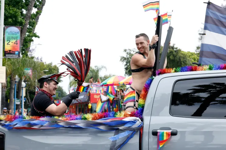 Một người đàn ông cầm flogger đánh người đang ông cười tươi dựa vào X-frame