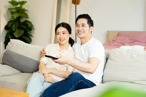 Người nam một tay cầm điều khiển tivi một tay ôm người nữ, hai người đang tươi cười trên sofa